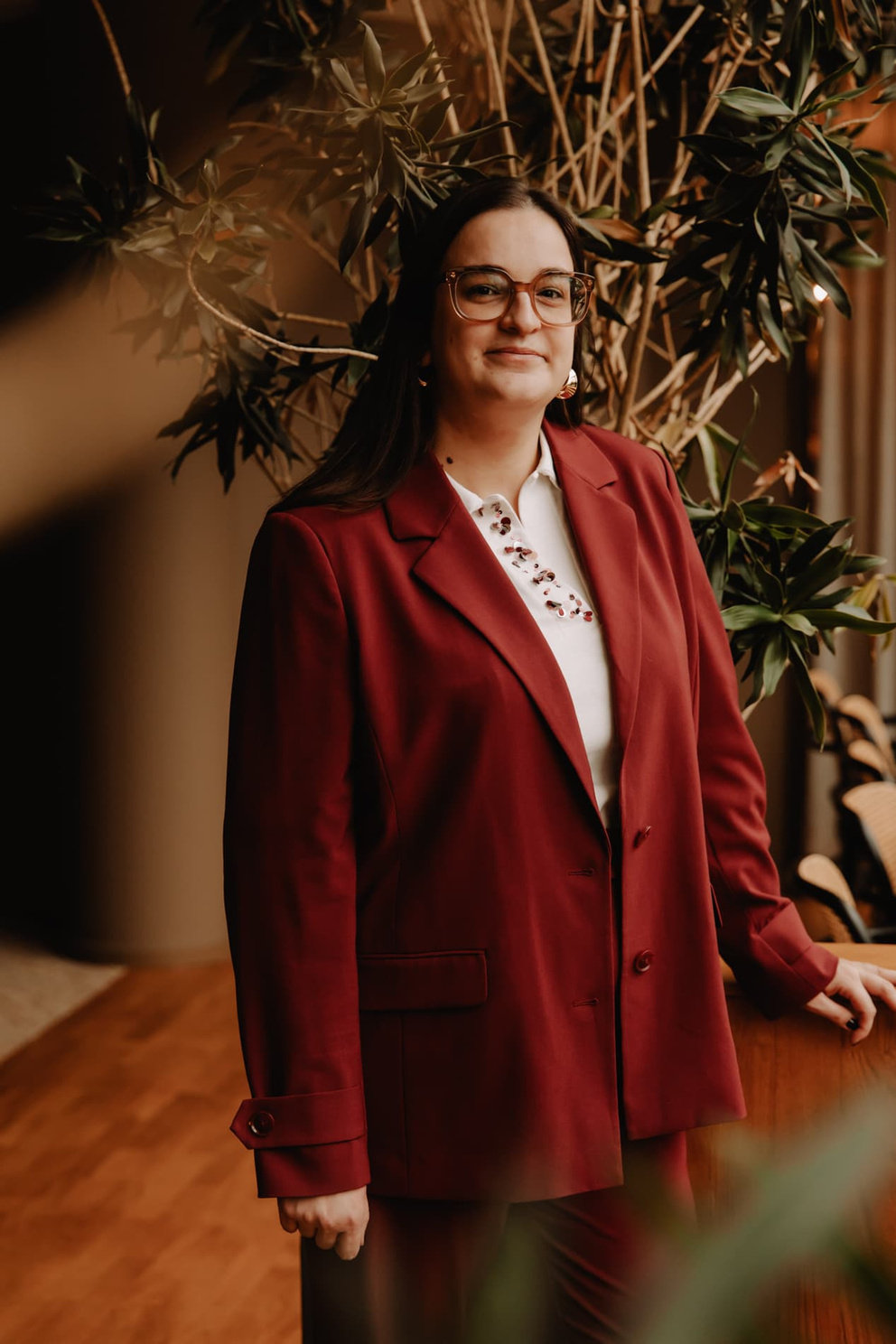 Vrouw in een rode blazer met een blouse, staand bij een groene plant in een moderne interieursetting.