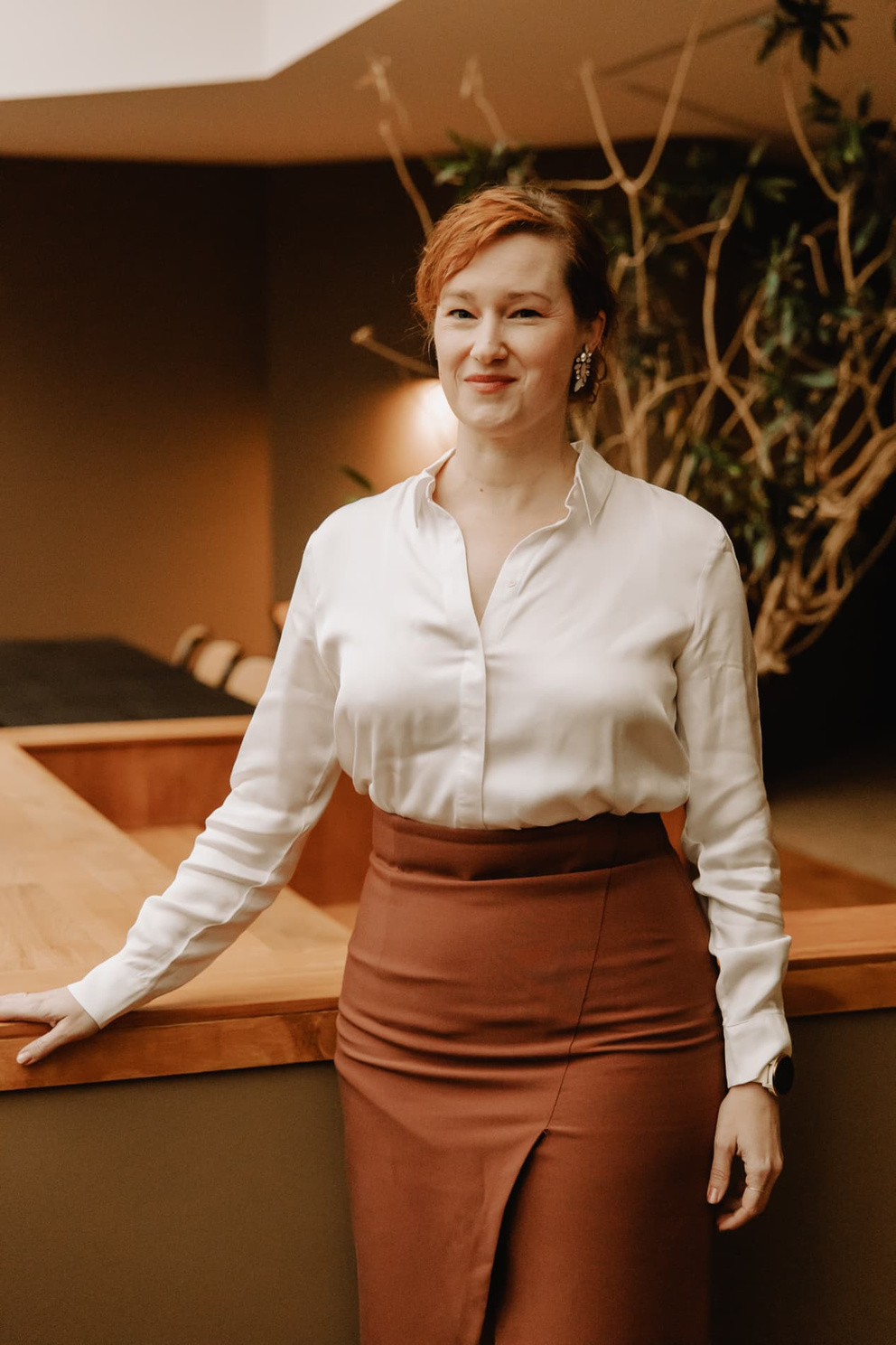 Vrouw in wit overhemd en bruine rok poseert zelfverzekerd bij een houten balie in een moderne, sfeervolle ruimte.