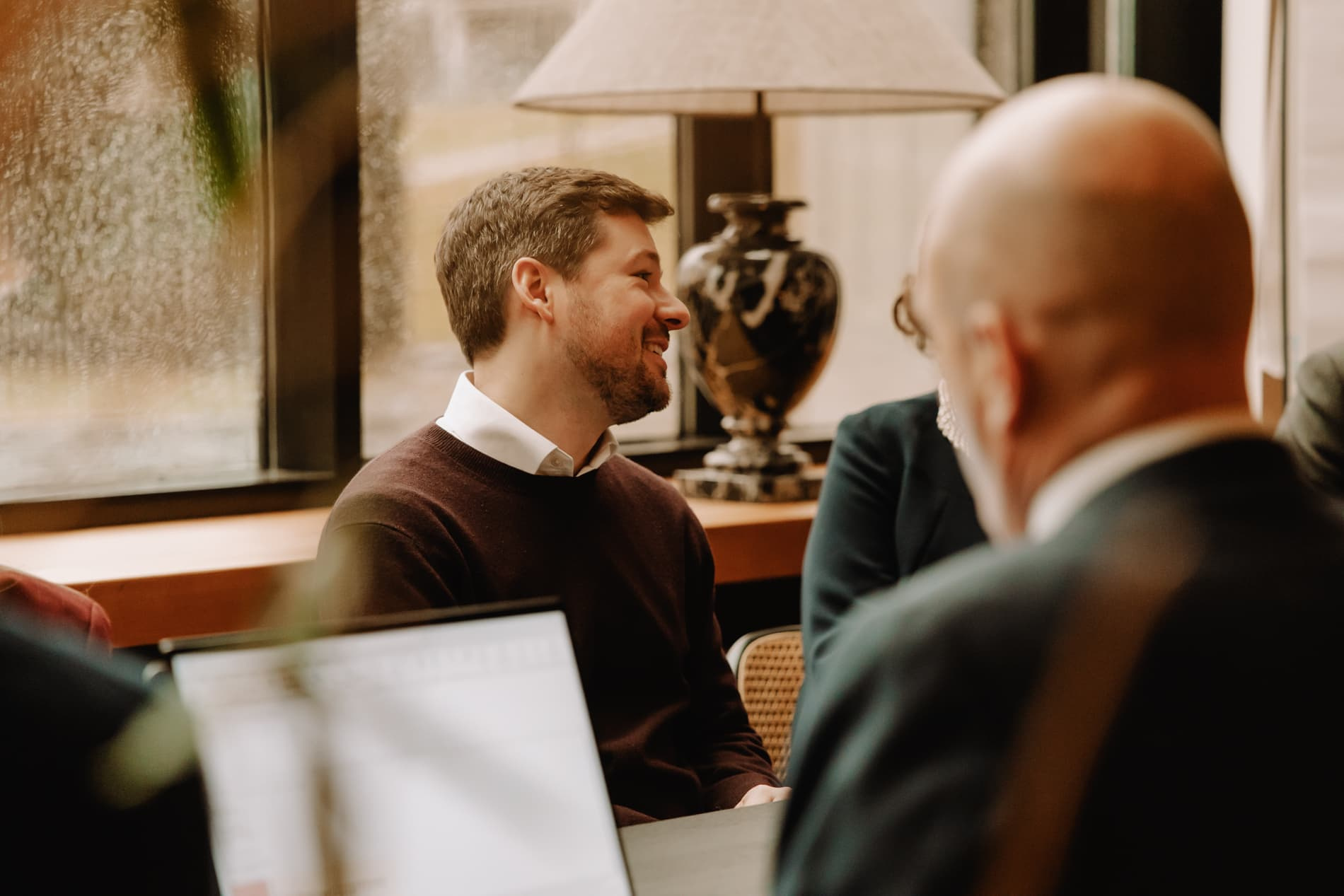 Een man in een donkere trui lacht tijdens een gesprek, met een lamp en een vaas op de achtergrond.