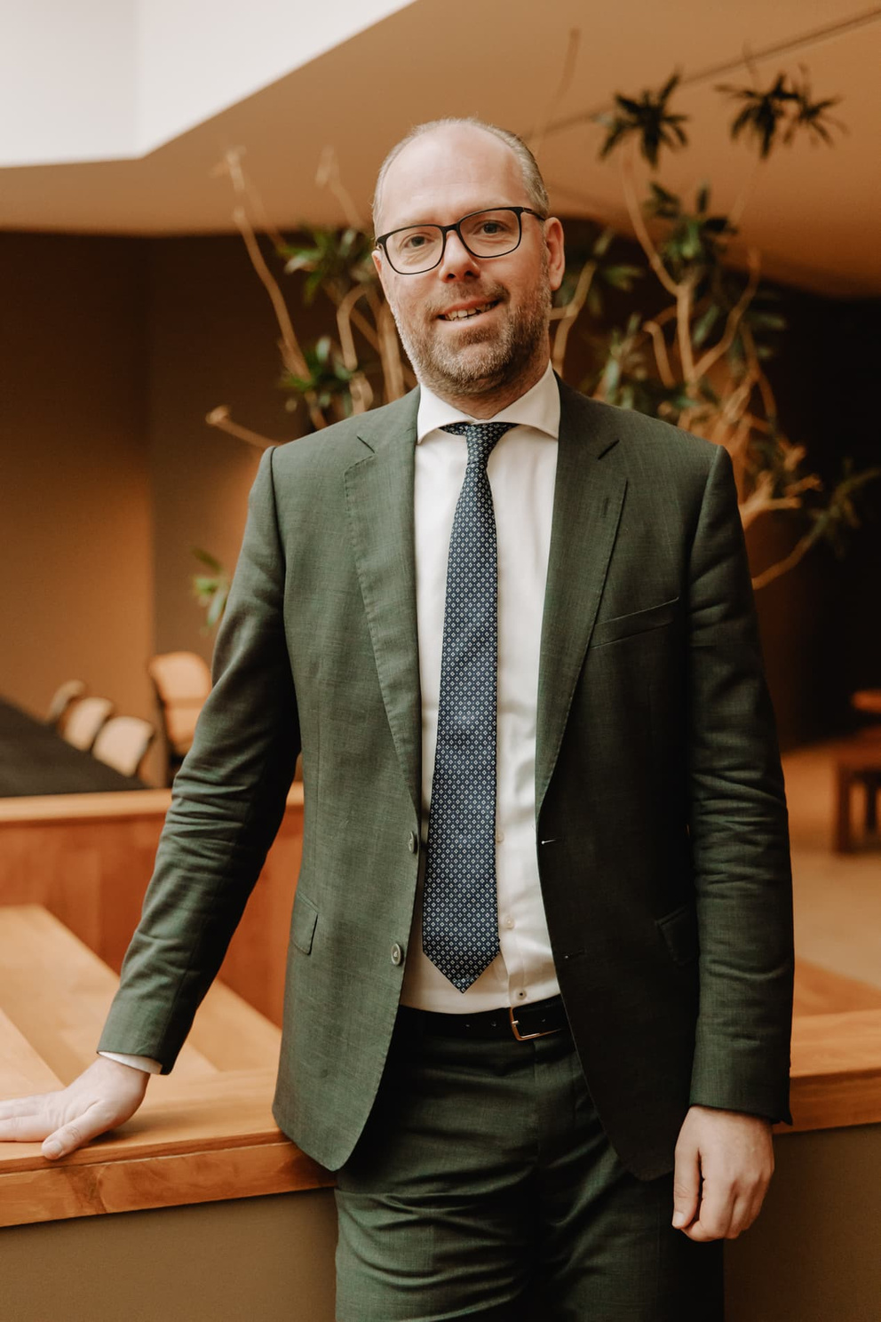 Man in een groen pak met een lichtgekleurde overhemd en stropdas, staande bij een houten balustrade in een moderne ruimte.