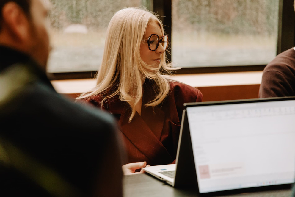 Blonde vrouw met lange haren en een bril, zittend aan een tafel met een laptop, kijkt attent naar iets buiten beeld.