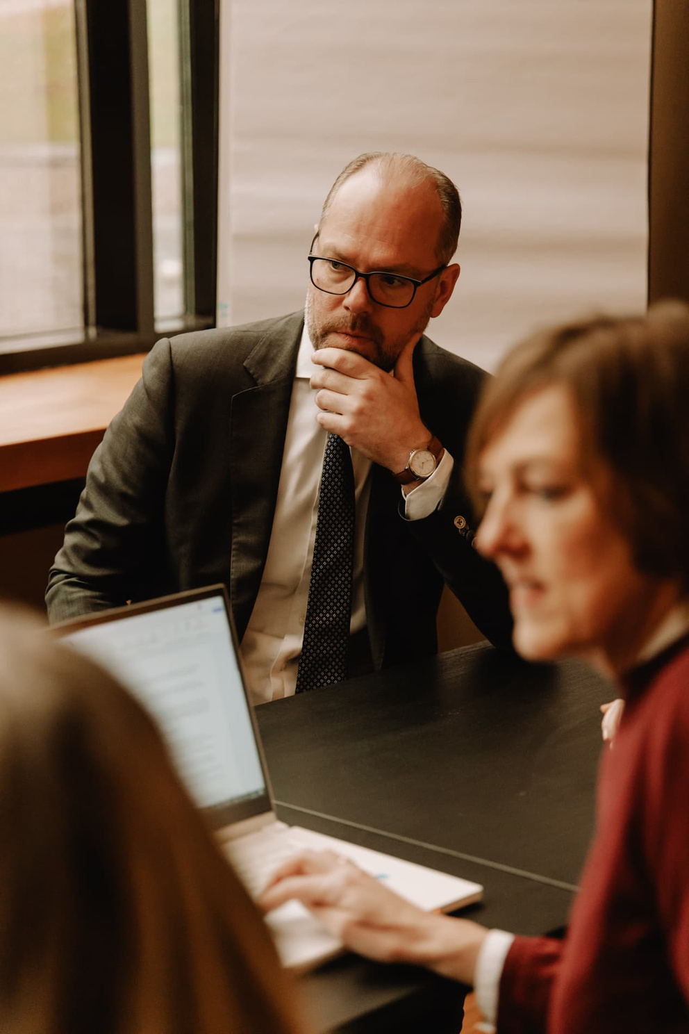 Een man in een pak luistert aandachtig naar een gesprek aan een vergadertafel, terwijl een vrouw aan de voorkant typt.