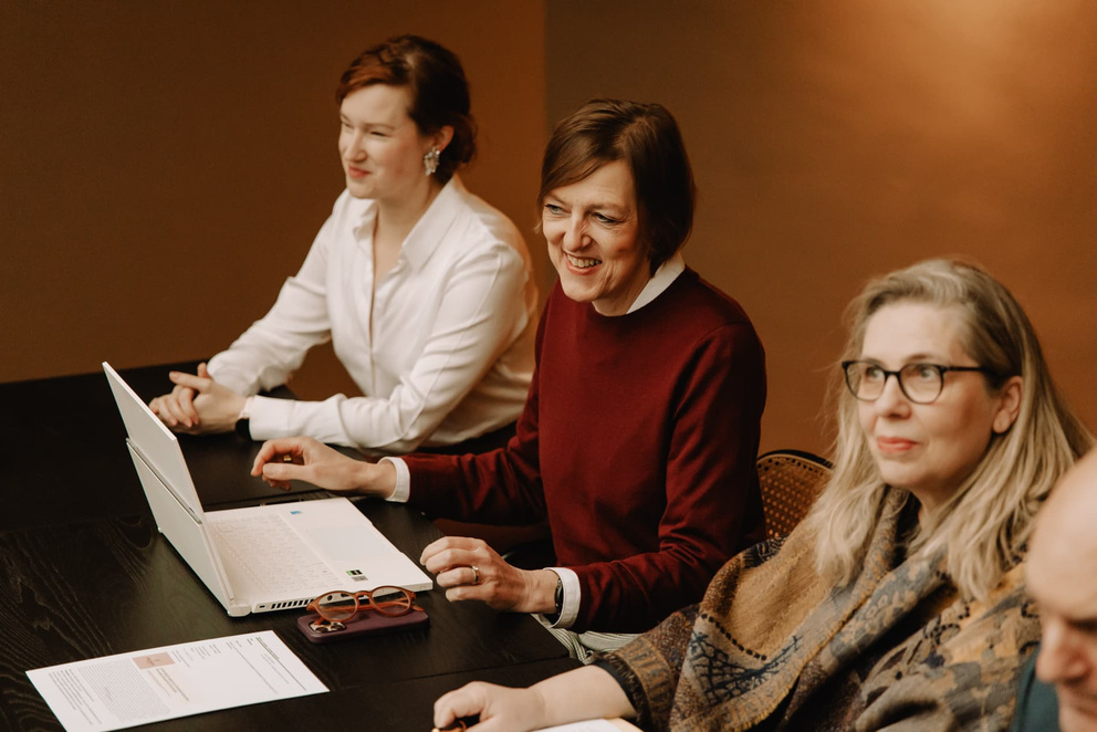 Vier vrouwen zitten aan een tafel, lachend en gefocust op een laptop, met documenten en notities voor zich.