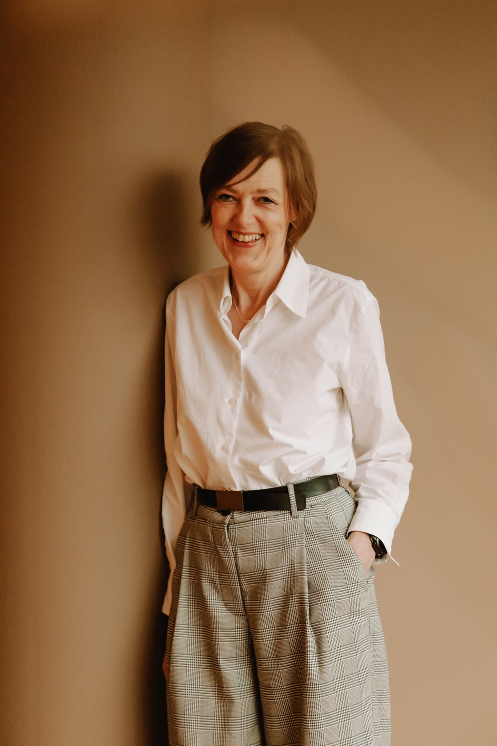 Vrouw in een witte blouse en geplooide broek, leunend tegen een muur en lachend naar de camera.