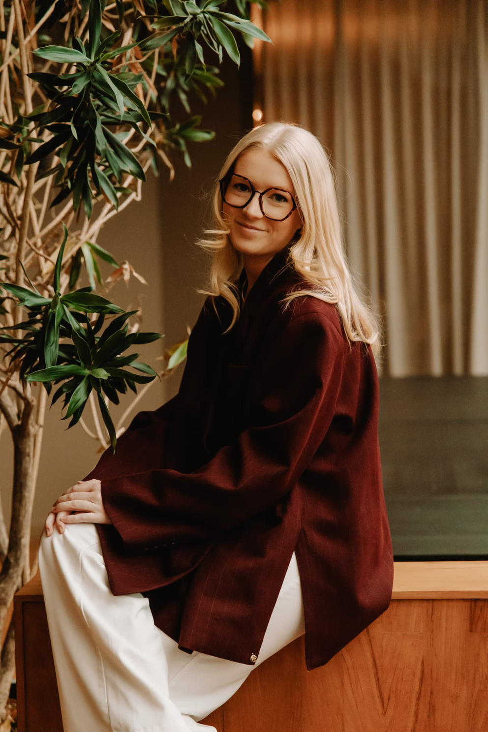 Jonge vrouw met lange, blonde haren en een paarse blouse zit op een houten bank, omringd door groene planten.