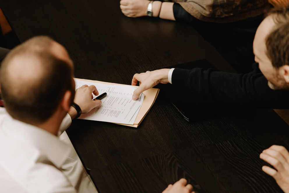 Mensen aan een vergadertafel bespreken documenten en notities. Eén persoon wijst op een papieren document.