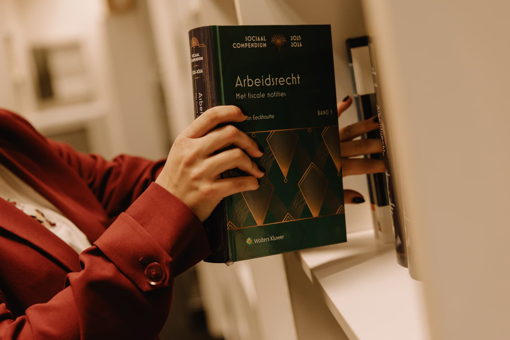 Een hand pakt een boek over arbeidsrecht uit een boekenkast, met een dieprode blazer zichtbaar.