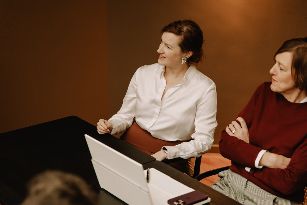 Twee vrouwen zitten aan een tafel, verdiept in gesprek. De een draagt een witte blouse, de ander een bordeauxkleurige trui.