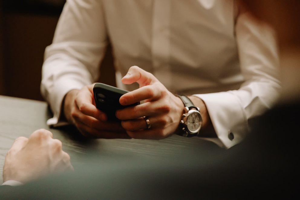 Een man in een wit shirt kijkt naar zijn smartphone terwijl hij aan een tafel zit, met een horloge om zijn pols.