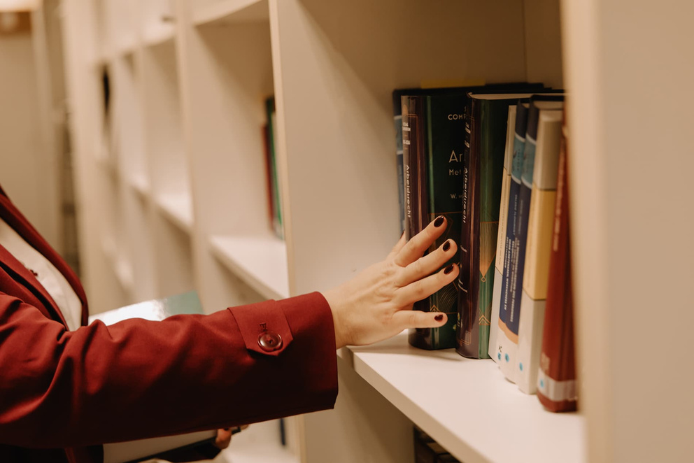 Een hand reikt naar boeken op een plankenwand in een bibliotheek of boekenkast.