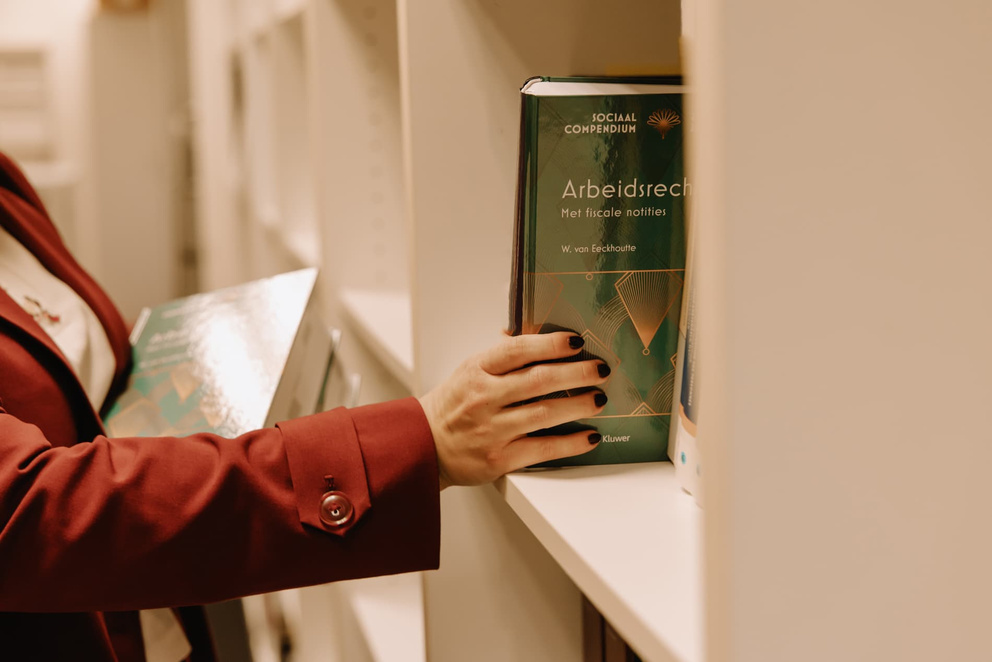 Een hand pakt een boek over arbeidsrecht uit een witte boekenkast.