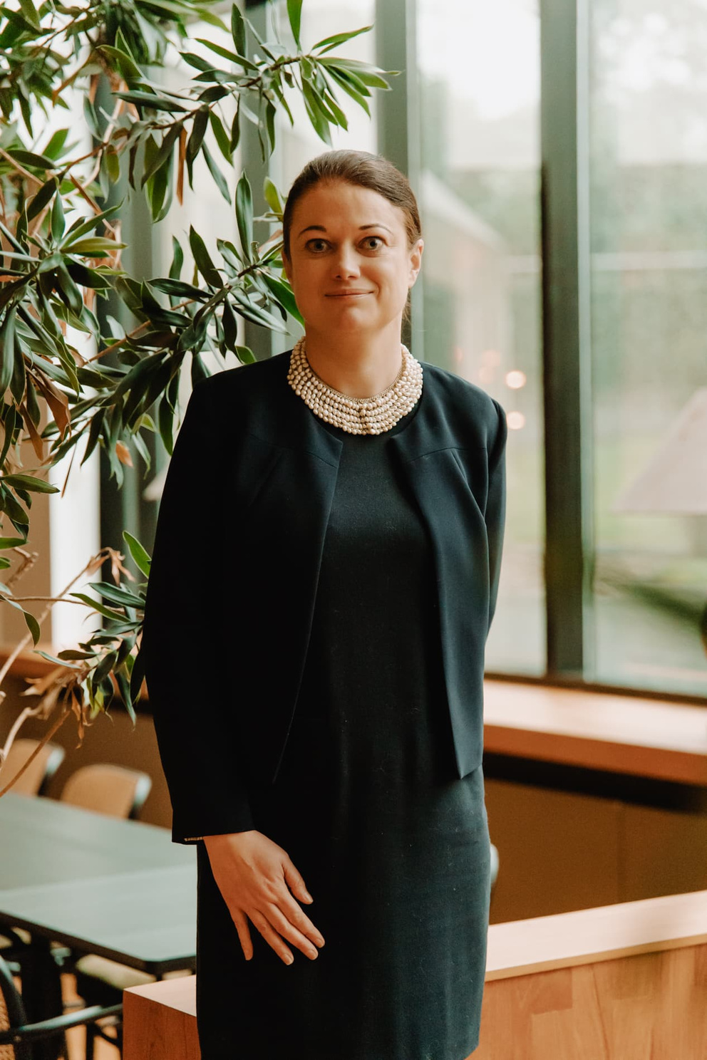 Vrouw in een zwarte outfit met een parelketting, staand bij een groot raam met planten op de achtergrond.