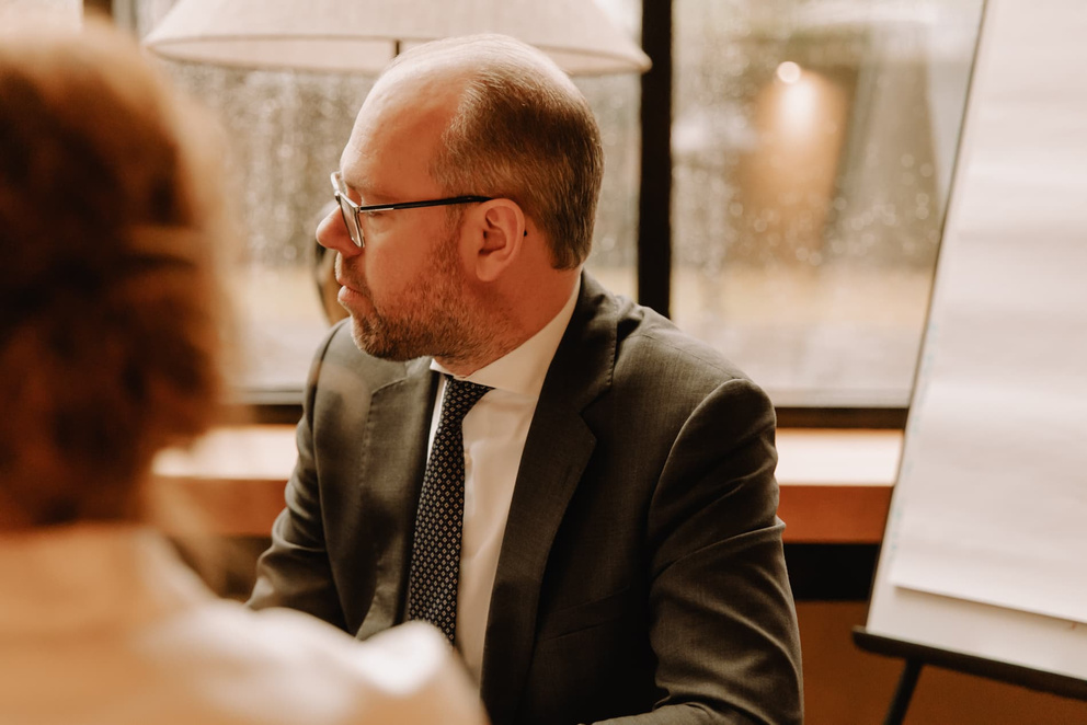 Twee zakelijke mensen in een kantooromgeving, met een focus op een man in een pak en een vrouw in het zicht.